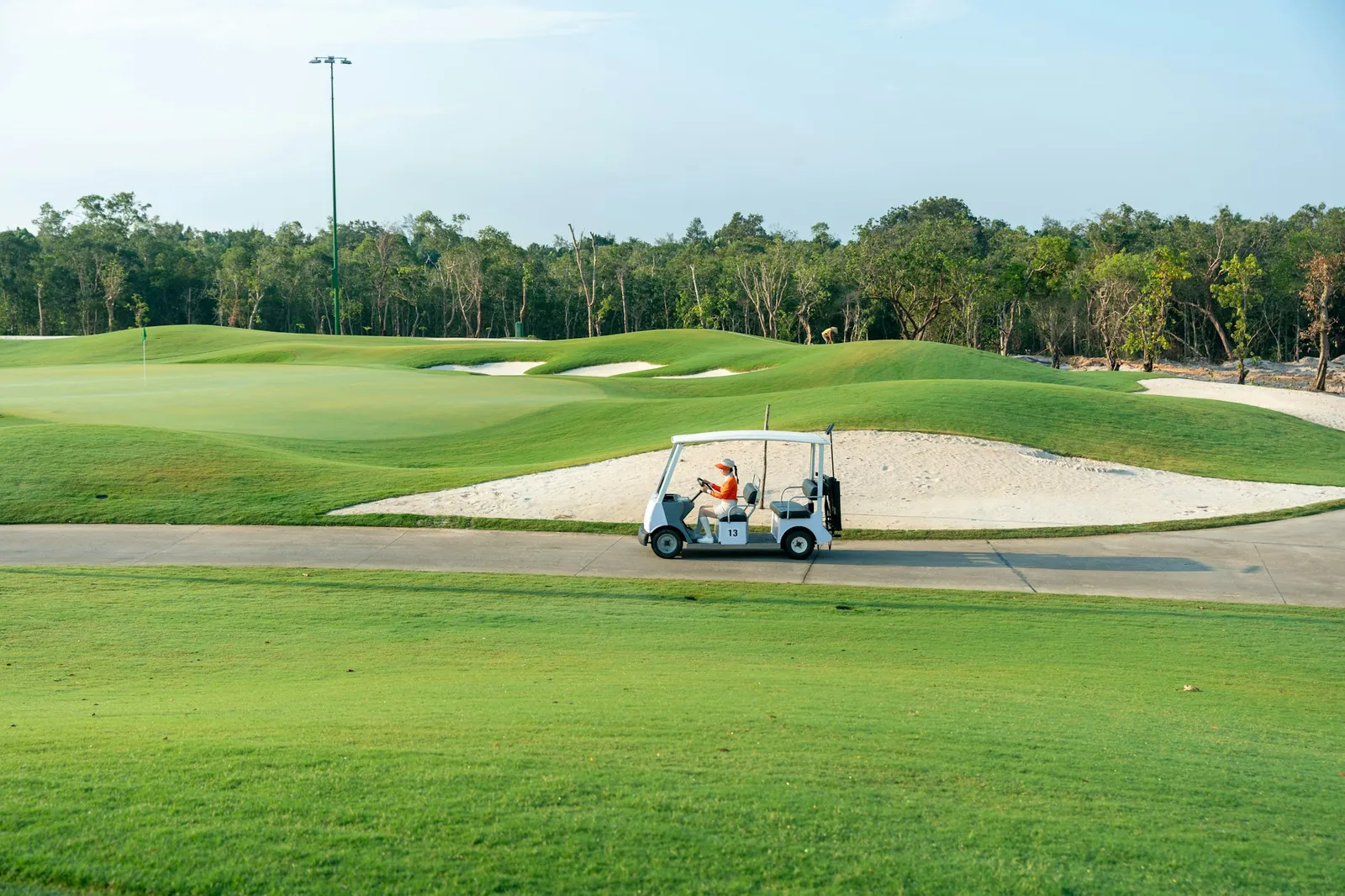 Golfbane med sandbunkere og en elbil på stien, omgitt av tropisk skog