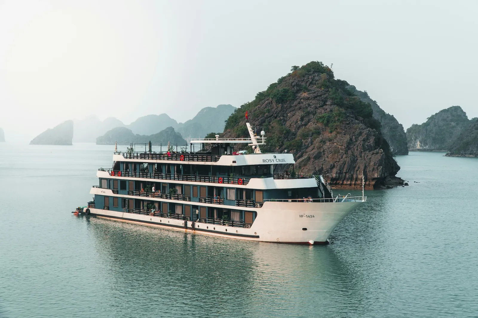 Cruiseskip i flere etasjer seilende mellom kalksteinsøyer i Ha Long-bukta i dis
