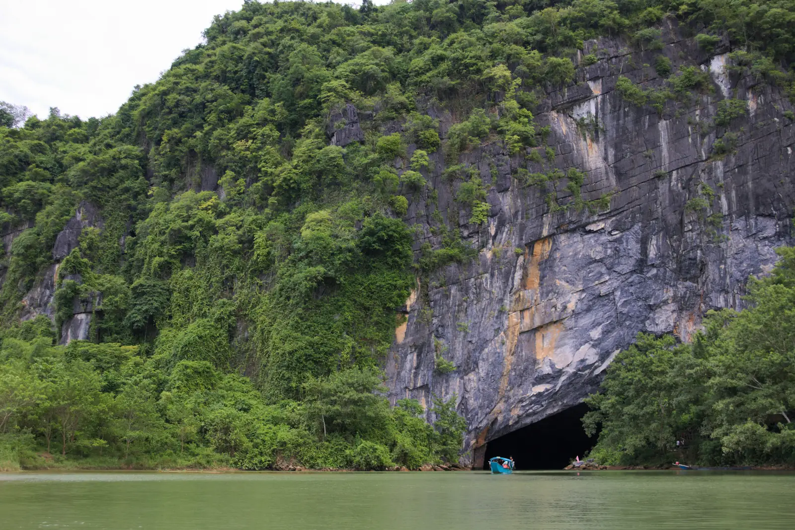 Inngangen til Phong Nha-grotten under en grønn kalksteinsvegg, med en liten båt på elva
