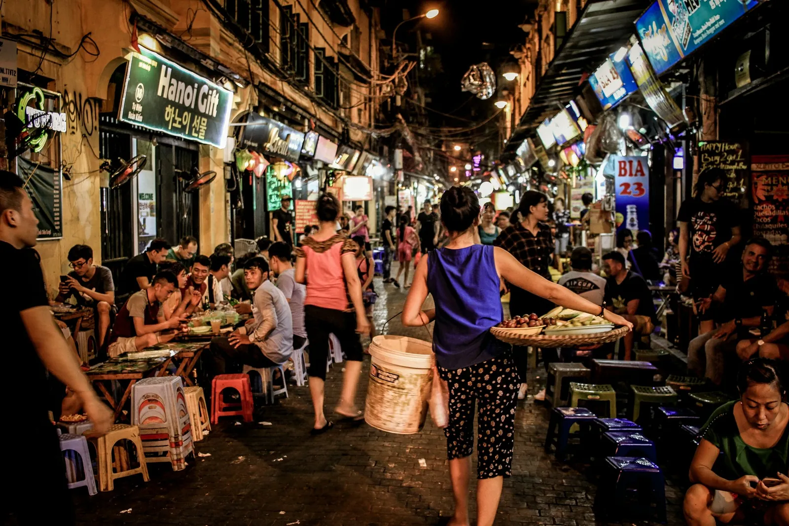 Folk på lave plastkrakker langs en trang gate fylt av barer og lys i Hanois gamleby om natten