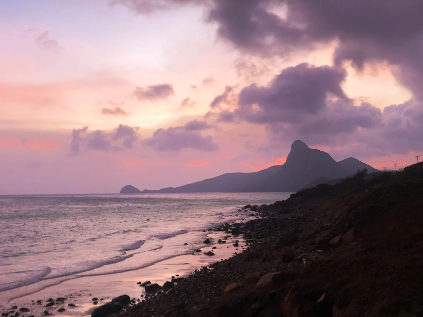 Rosa solnedgang over steinstrand og fjellsilhuetten på Con Dao-øya