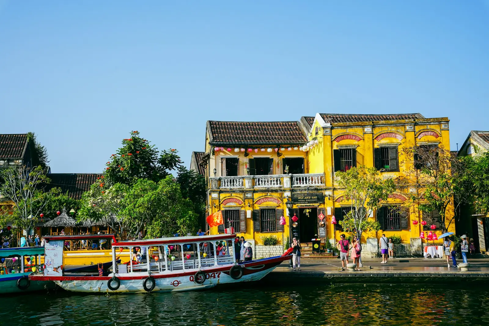 Gule hus og en turistbåt langs elvebredden i Hoi An